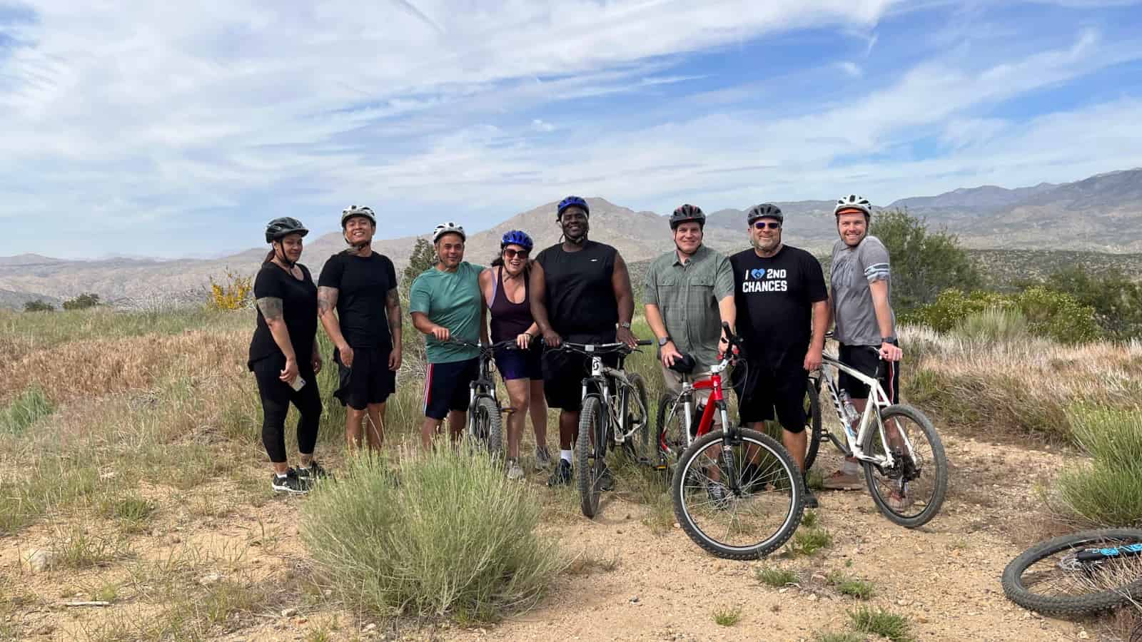 Eight smiling adults of various skin colors, body types, and genders, all dressed in athletic gear or general outdoor clothing and wearing bicycle safety helmets. They are standing in an informal row in the middle of a rocky arid landscape with low bushes and dried grass, and the sky is blue but filled with thin, windswept white clouds. Three of the people are holding up mountain bikes, and there is the front wheel of another mountain bike on its side in the near right foreground. In the far distance behind the group are low hills.