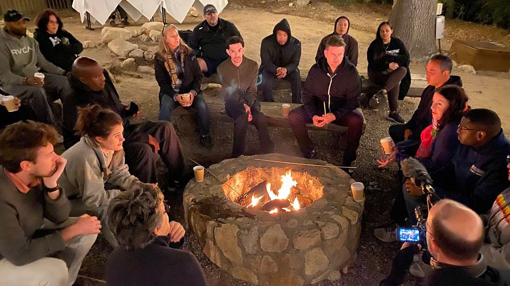 A lit fire burns in a rocky fire pit in a dusty brown park environment at night. Seventeen adults of various ages, skin colors, and genders sit on rough wooden benches in a loose semi-circle around the fire pit. A woman on the right side of the fire is speaking, and others in the semicircle are looking at her and listening. In the bottom right corner, a man holding a video camera with attached boom mic is recording the scene.