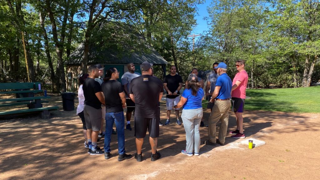 Twelve adults of various skin colors and genders stand in a circle in the partial shade in a park on a sunny day. They are all holding onto a loop of rope at waist level as part of a team-building exercise.