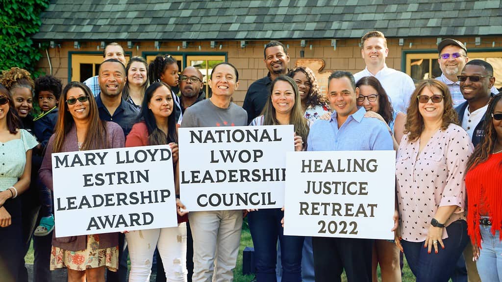 A large group of people of various ages, skin colors, and genders are standing outside in front of a building with a low green roof. They are smiling at the camera and holding up three signs, which read in order from left to right, MARY LLOYD ESTRIN LEADERSHIP AWARD, NATIONAL L.W.O.P. LEADERSHIP COUNCIL, and  HEALING JUSTICE RETREAT 2022.