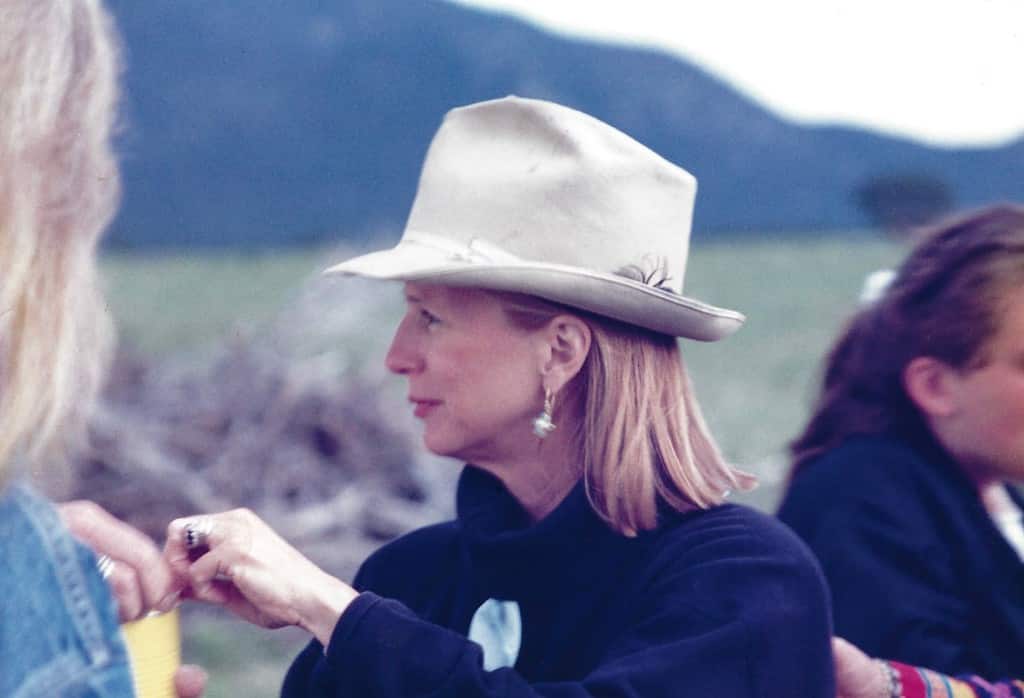 Mary Lloyd Estrin, a middle-aged caucasian woman with shoulder-length straight blonde hair, is standing outside, looking off to her right (our left) with her face in profile. She is wearing a large white  brimmed hat and has a gold and white earring that hangs down, and she is wearing a dark blue pullover. Other people are around her, but not in focus and mostly out of frame. In the far distance are softly blurred silhouettes of hills or low mountains.