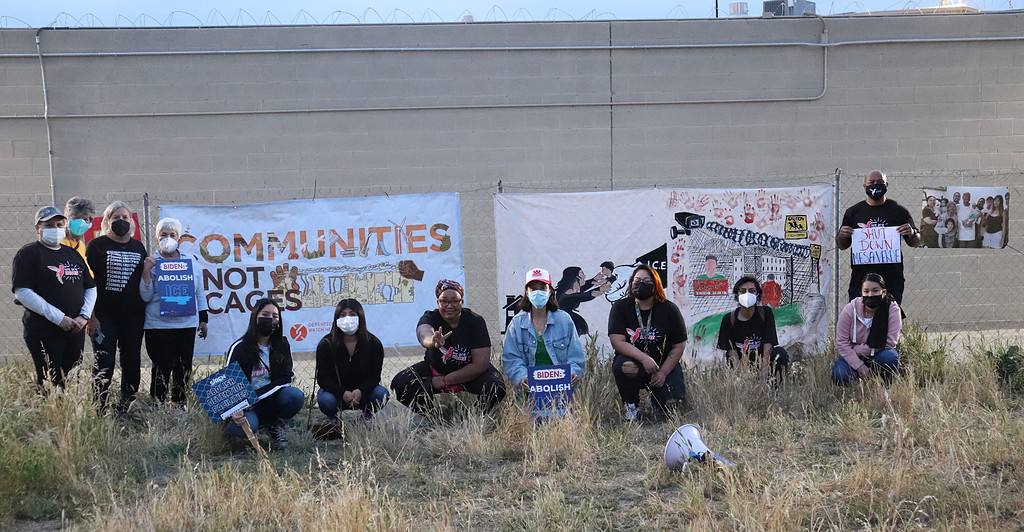 A group of protestors of various genders, ages, and skin colors, most wearing COVID-era masks, stand in front of a 20-foot-tall, windowless, beige cinder-block building topped by razor wire. A chain-link fence stands between the building and the protestors, and the protestors have hung two large banners on the fence. The left banner reads "COMMUNITIES NOT CAGES," and the right banner has two images: on the left side, a woman reaches out for a child who is gripped in an oversized hand with ICE written on the sleeve; on the right side, two men look out from the windows of a prison, underneath a sky made out of red hand prints.