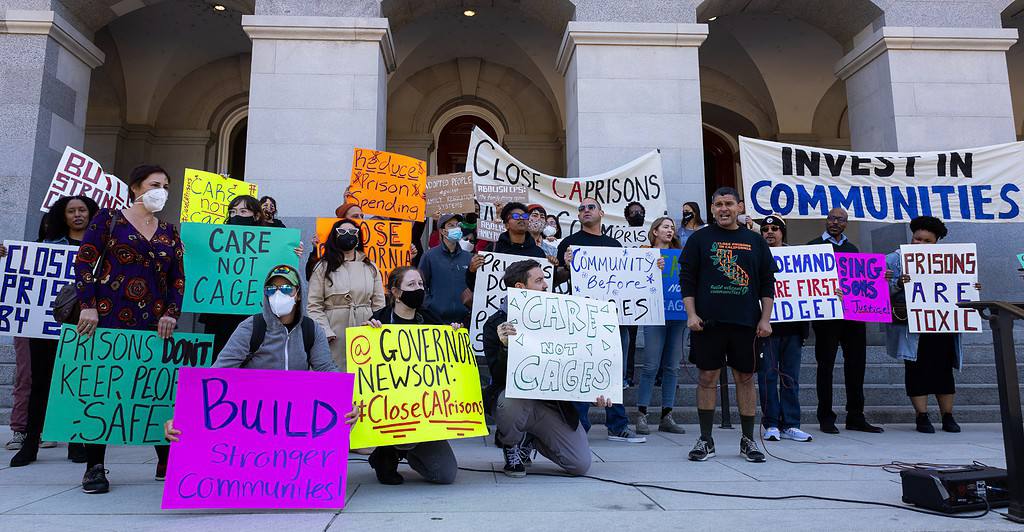 A group of young people wearing COVID-era face masks are gathered before a municipal building. Most are standing, but three people in the foreground are kneeling. Most of the protestors are holding homemade signs with messages such as, PRISONS DON'T KEEP PEOPLE SAFE, CARE NOT CAGES, INVEST IN COMMUNITIES, and PRISONS ARE TOXIC.
